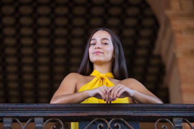 Genç bir kadın, esmer, Latin, sarı bir gömlek giyiyor, kameraya bakıyor, mutlu ve gülümsüyor, Seville 'deki İspanya Meydanı' nda metal bir korkuluğa yaslanıyor. Kavram: Gençlik, seyahat, hedefler.