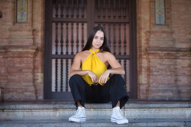 Genç bir kadın, esmer, Latin, sarı gömlek, siyah kot pantolon ve beyaz spor ayakkabılar giyiyor, güçlü, etkileyici bir bakışla, bazı basamaklara oturuyor. Kavram: gençlik, moda, moda, modern.