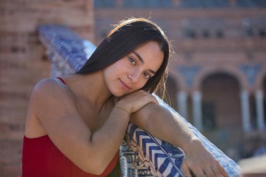 Genç, koyu renk saçlı, Latin bir kadının portresi kırmızı bir elbise giymiş, tatlı ve narin, yüzü kolunda dinlenen ve fayanslı bir korkuluğa yaslanmış kameraya bakıyor. Kavram: bakışlar, şefkat.