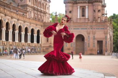 Genç, güzel, sarışın, kırmızı flamenko elbiseli, saçında çiçekler ve altın mücevherlerle İspanya meydanında flamenko pozları veriyor. Dans konsepti, sanat, tipik İspanyol..