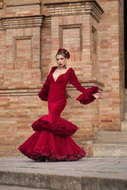 Genç, güzel, sarışın, kırmızı flamenko elbiseli, saçında çiçekler ve altın mücevherlerle İspanya meydanında flamenko dansı yapan bir kadın. Dans konsepti, sanat. Dikey konum.