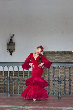 Genç, güzel, sarışın, kırmızı flamenko elbiseli, saçında çiçekler ve altın mücevherlerle, Seville 'de İspanya meydanında metal bir korkuluğa yaslanmış flamenko pozları veriyor. Dikey konum.