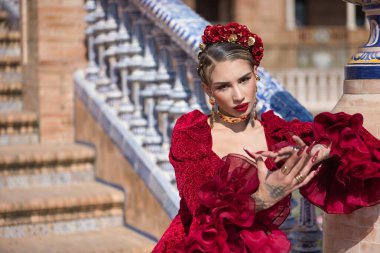 Genç, güzel, sarışın bir kadının portresi, kırmızı flamenko elbisesi, saçında çiçekler ve altın mücevherler, Sevilla 'da İspanyol meydanında flamenko pozları veriyor. Dans konsepti, sanat