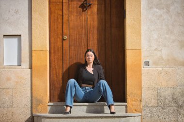 Genç bir kadın, çekici, esmer, mavi gözlü, İspanyol, siyah ceket ve bluz giyiyor, kot pantolon giyiyor, kameraya bakıyor, kapı eşiğinde oturuyor. Konsept: model, güzellik, moda, moda.