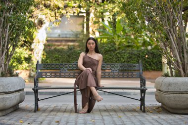 Genç bir kadın, güzel, esmer, mavi gözlü, İspanyol, zarif kahverengi bir şifon elbise ve altın topuklu ayakkabı giyiyor, kentteki bir bankta otururken poz veriyor. Konsept: model, güzellik, moda, moda.