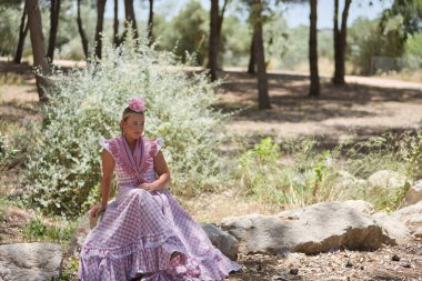 Tipik Endülüs flamenko kostümlü genç, güzel, sarışın bir kadın tarlada bir taşın üzerinde dinlenen küçük şalla ve pembe çiçekle hacca gidecek. Bölgesel kostümler, tipik.