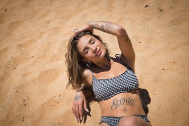 Genç, güzel, Latin bir kadının portresi dövmeli, siyah beyaz kareli bikini giyiyor, kumsalda gözleri kapalı, güneşleniyor. Konsept: tatil, yaz, güneş.