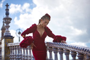 Genç, güzel, kırmızı flamenko elbiseli sarışın kadın, saçında çiçekler ve altın mücevherler, İspanya 'nın Seville meydanındaki güzel bir köprüde flamenko pozları veriyor. Dans konsepti, sanat, tipik.