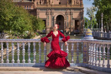 Genç, güzel, sarışın, kırmızı flamenko elbiseli, saçında çiçekler ve altın mücevherlerle, Seville 'de İspanya meydanında fayanslı bir korkuluğa yaslanmış flamenko pozları veriyor. Dans konsepti, sanat.