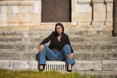 Genç bir kadın, çekici, esmer, mavi gözlü, İspanyol, siyah ceket giyen, kot giyen, topuklu giyen, şehir merkezinde merdivenlerde otururken poz veren. Konsept: model, güzellik, moda, moda.