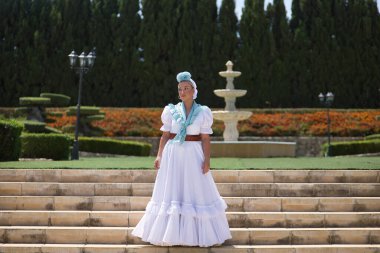 Genç, güzel, sarışın kadın tipik Endülüs flamenko kostümlü açık mavi şallı ve çiçekli hac için merdivenlerde poz veriyor. Konsept bölgesel kostümler, tipik, İspanyolca.
