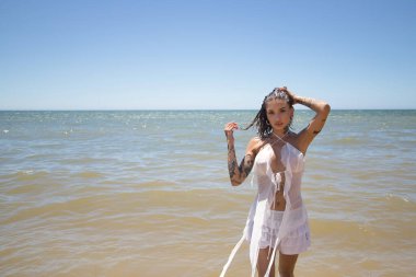 Genç, güzel, dövmeli Latin bir kadın, beyaz bir elbise giyiyor, denizden şık ve cazibeli bir şekilde çıkıyor, bir eli kafasında, güneşli bir plaj gününde. Konsept: tatil, deniz, sahil.