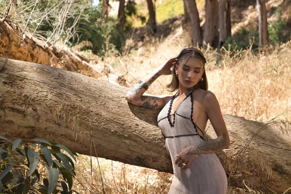 Genç, güzel, dövmeli bir Latin kadın, beyaz bir örgü örüyor, melankolik ve üzgün görünüyor, yere bakıyor, bir ağaç gövdesine yaslanıyor. Kavram: kadınlık, yalnızlık, doğa.