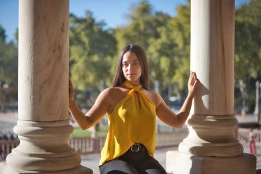 Genç bir kadın, esmer, Latin, sarı tişörtlü, iki mermer sütunun arasında oturuyor, dinleniyor, İspanya meydanında, Seville 'de. Kavram: gençlik, turizm, seyahat, büyüleyici yerler.