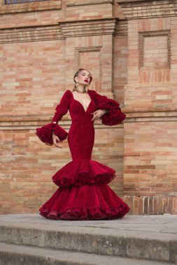 Genç, güzel, sarışın, kırmızı flamenko elbiseli, saçında çiçekler ve altın mücevherlerle İspanya meydanında flamenko dansı yapan bir kadın. Dans konsepti, sanat. Dikey konum.