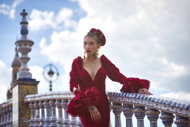 Genç, güzel, kırmızı flamenko elbiseli sarışın kadın, saçında çiçekler ve altın mücevherler, İspanya 'nın Seville meydanındaki güzel bir köprüde flamenko pozları veriyor. Dans konsepti, sanat, tipik.