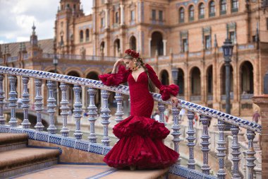 Genç, güzel, kırmızı flamenko elbiseli sarışın kadın, saçında çiçekler ve altın mücevherler, İspanya 'nın Seville meydanındaki güzel bir köprüde flamenko pozları veriyor. Dans konsepti, sanat, tipik.