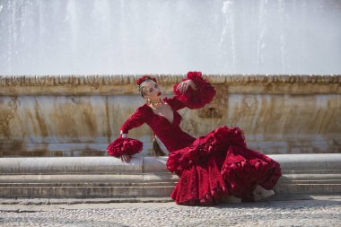 Genç, güzel, sarışın, kırmızı flamenko elbiseli, saçında çiçekler ve altın mücevherlerle, İspanya 'nın Seville meydanındaki güzel bir çeşmede flamenko pozları veriyor. Dans konsepti, sanat.
