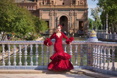 Genç, güzel, sarışın, kırmızı flamenko elbiseli, saçında çiçekler ve altın mücevherlerle, Seville 'de İspanya meydanında fayanslı bir korkuluğa yaslanmış flamenko pozları veriyor. Dans konsepti, sanat.