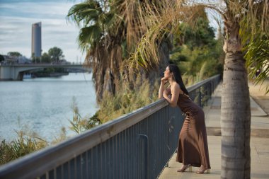 Genç bir kadın, güzel, esmer, mavi gözlü, İspanyol, zarif kahverengi bir şifon elbise ve altın topuklu ayakkabı giyiyor, eli çenesinde, mesafeye bakıyor, metal bir korkuluğa yaslanıyor..