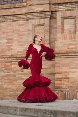 Genç, güzel, sarışın, kırmızı flamenko elbiseli, saçında çiçekler ve altın mücevherlerle İspanya meydanında flamenko dansı yapan bir kadın. Dans konsepti, sanat. Dikey konum.