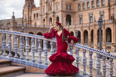 Genç, güzel, kırmızı flamenko elbiseli sarışın kadın, saçında çiçekler ve altın mücevherler, İspanya 'nın Seville meydanındaki güzel bir köprüde flamenko pozları veriyor. Dans konsepti, sanat, tipik.