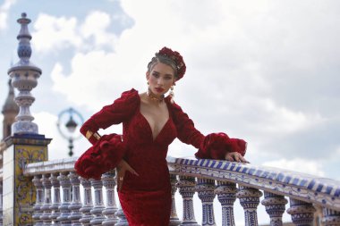 Genç, güzel, kırmızı flamenko elbiseli sarışın kadın, saçında çiçekler ve altın mücevherler, İspanya 'nın Seville meydanındaki güzel bir köprüde flamenko pozları veriyor. Dans konsepti, sanat, tipik.