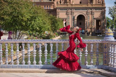 Genç, güzel, sarışın, kırmızı flamenko elbiseli, saçında çiçekler ve altın mücevherlerle, Seville 'de İspanya meydanında fayanslı bir korkuluğa yaslanmış flamenko pozları veriyor. Dans konsepti, sanat.