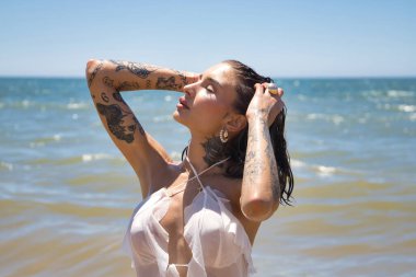 Genç, güzel, Latin bir kadının portresi, dövmeli, beyaz bir elbise giyiyor, ıslak, elleri başının üstünde, güneşli bir sahil gününde şık ve görkemli bir şekilde denizde poz veriyor. Konsept: tatil.