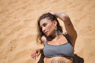 Siyah beyaz kareli bikini giyen, kumsalda güneşlenirken dikkati dağılmış genç, güzel, Latin bir kadının portresi. Konsept: tatil, yaz.