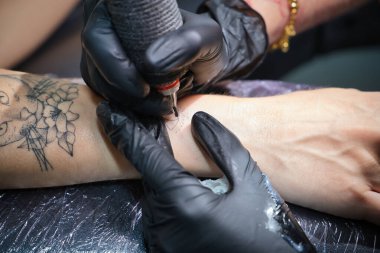 Siyah eldiven giyen, dövme makinesi tutan, stüdyosunda başka bir kadının koluna dövme yapan bir kadın dövme sanatçısının ellerinin ayrıntıları. Kavram: mürekkep, iğne, dövme, tasarım, bakım.