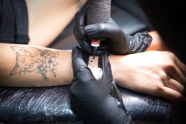 Siyah eldiven giyen, dövme makinesi tutan, stüdyosunda başka bir kadının koluna dövme yapan bir kadın dövme sanatçısının ellerinin ayrıntıları. Kavram: mürekkep, iğne, dövme, tasarım, bakım.