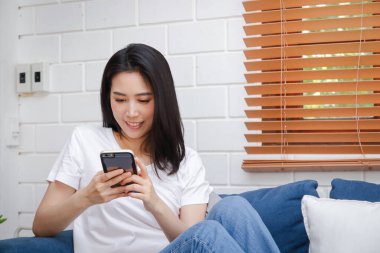 Akıllı telefon tutan Asyalı genç bir kadın sosyal medyada oynuyor, evdeki oturma odasındaki kanepede oturuyor. İletişim teknolojisi kavramı, modern iş yapma.