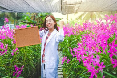 Bitki genetiği araştırmacısı, orkide bahçesinde beyaz bir elbise giyiyor. Elinde bir tabela var. Mesaj yazabilir.