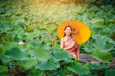Antik Tayland kostümleri giymiş Asyalı kızlar tahta bir teknede oturuyorlar. Lotus havuzunda sarı bir şemsiye tutuyorlar.