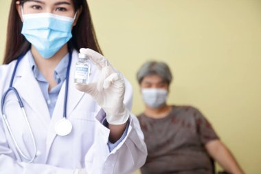 Mavi maske takan genç ve güzel bir Asyalı doktor bir şişe koronavirüs aşısını (COVID-19) hasta birinin arkasında tutuyor. Hastane sağlık hizmeti konsepti. Antiviral aşı