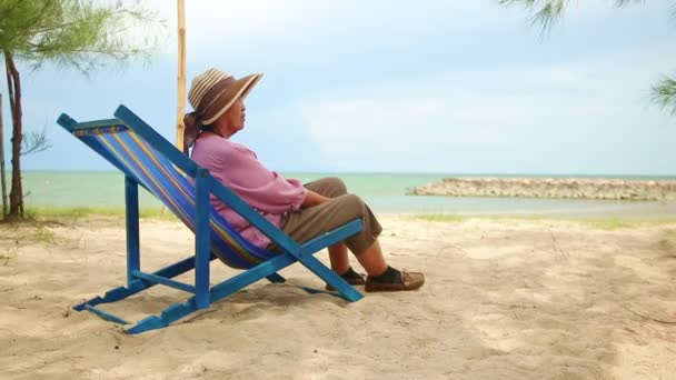 一位戴着帽子的亚洲老妇人坐在海滩旁边的躺椅上 老年人在退休年龄旅行性质的概念 复制空间 — 图库视频影像