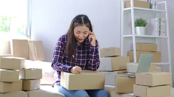 若いアジアの女性はインターネットを通してオンラインで販売するビジネスをしている 郵便で小包を送る準備をするために顧客に話すためにスマートフォンを保持します オンライン販売の概念 — ストック動画