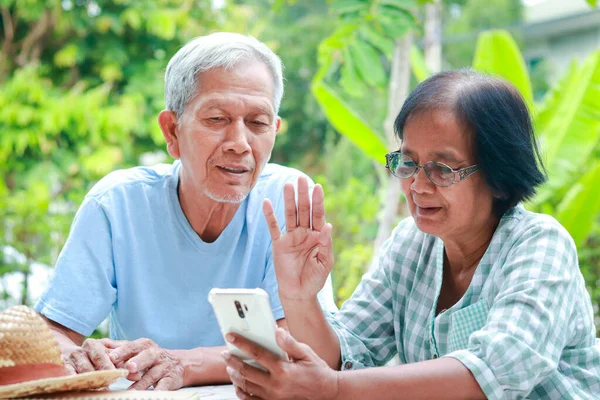 Asian elderly couple happy living at home Sitting in the garden holding a smartphone, chatting online with your child. Family concept, health care for the elderly in retirement age