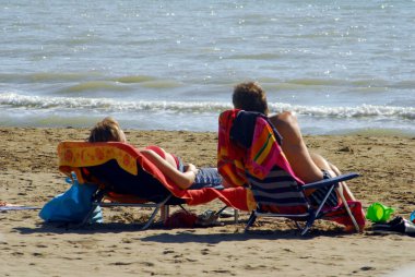 Yaz tatilinde kumlu bir sahilde güverte sandalyesinde uzanan erkek ve kadın.