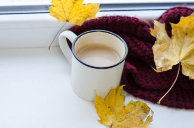 Pencerenin yanında bir fincan kahve eşarp ve sarı sonbahar yapraklarıyla