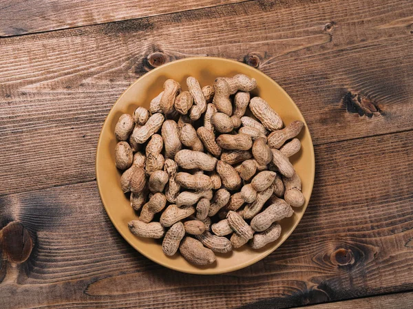 Freshly roasted peanuts on a yellow ceramic plate on a rough wooden tabletop.