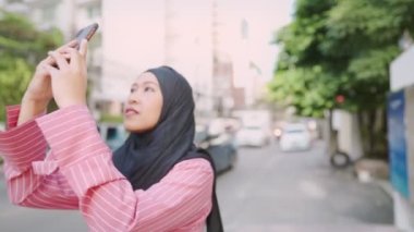 Asyalı müslüman bir kadın siyah başörtüsü takıyor. Güneşli bir günde, kalabalık bir yol kenarında modern bir şehir manzarası fotoğrafı çekiyor. Asya seyahati ve anıları, insanlar ve kullanışlı kablosuz teknoloji, modern teknoloji ve seyahat anıları.