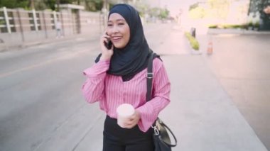 Asyalı Müslüman çalışan kadın iş yerine yürürken bir telefon görüşmesi yapıyor, kahve fincanı tutuyor, şehir merkezindeki sokak kenarında kahve molası veriyor, taşınabilir mobil cihaz, telefon görüşmesi, hafta sonları dinlendirici aktivite, basitçe yaşam tarzı.