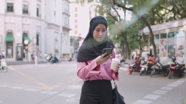 Asyalı genç Müslüman bir kadın telefon görüşmesi yapıyor, kahve molasında elinde kahve fincanı, taşınabilir mobil cihaz, Asya 'nın işlek şehir sahnesi, taksi çağırma servisi, kablosuz internet bağlantısı, modern din hayatı.