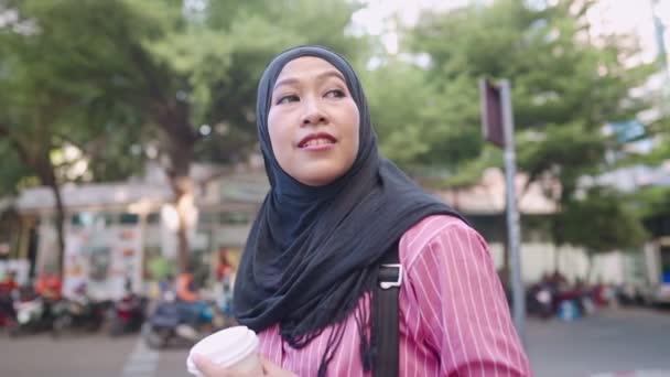 若いアジアのイスラム教徒の女性は 道路脇の歩行者の交差点に立って黒いヒジャーブを身に着けている 観光客の放浪は 都市の近代的な生活 車やバイクをバックグラウンドで通過見て楽しむ 無線インターネット接続 現代の宗教生活 — ストック動画