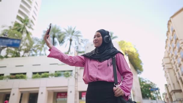 美しい建物のランドマークの前の道端に立つ若いイスラム教徒の女性は ヒジャーブを身に着けています自分で自撮り ライフスタイルと旅行 物語を共有します アジアの観光客 ワイヤレスインターネット接続 現代の宗教生活 — ストック動画
