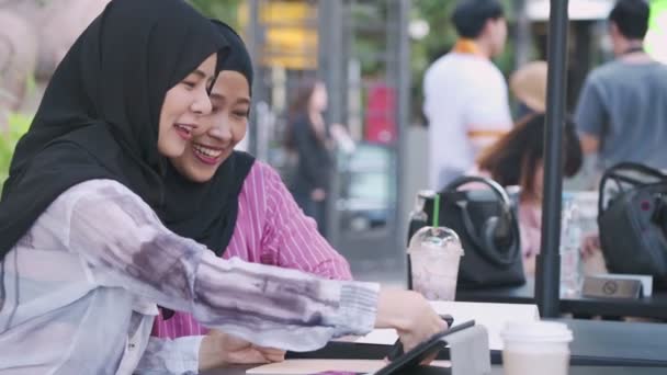 身穿头巾的穆斯林妇女的衣服在下午休息的时候在户外街道咖啡馆笑 两个笑容可亲的漂亮姑娘边坐着边看智能手机 玩得开心 无线上网 过着现代宗教生活 — 图库视频影像