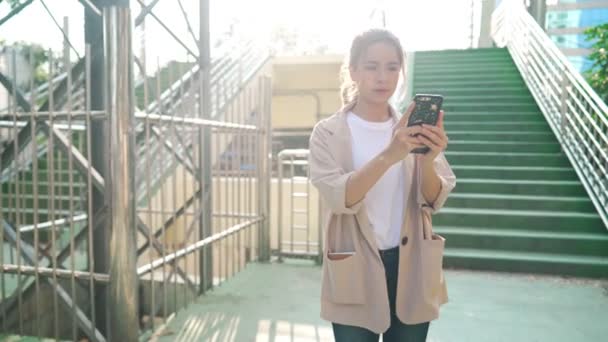 穿着休闲装的迷人女子手拿着智能手机在美丽的城市天桥上漫步 迷茫的年轻学生凝视着朋友送给她的方向 生活在智能技术 无线移动技术 网络连接中 — 图库视频影像