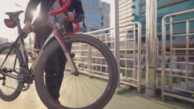 Güneş gözlüklü ve takım elbiseli Asyalı yakışıklı genç işadamı dışarıda yürüyor ve bisiklet taşıyor, arka planda modern cam bina var. Şehir üst geçidinde çarklar dönüyor, hobi ve şehir yaşam tarzı, çevresel koruma ya da sorunlar, insanların hobisi